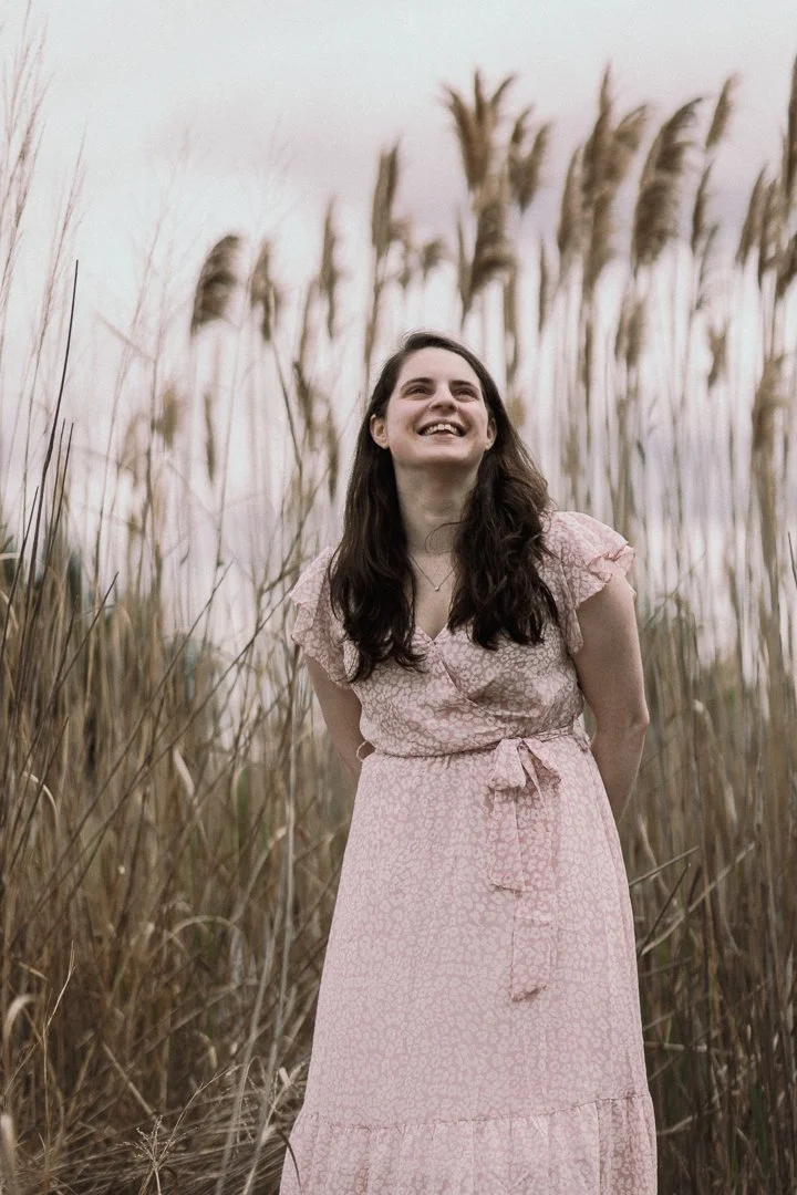 A woman with long dark hair smiling and standing in a field of tall grass at sunset, wearing a pink dress with short sleeves.
