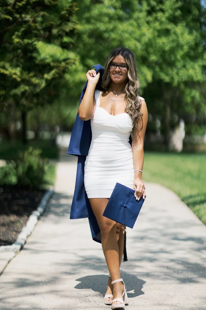 A young woman in a white dress and glasses, holding a graduation cap, walking on a sidewalk in a park, smiling.