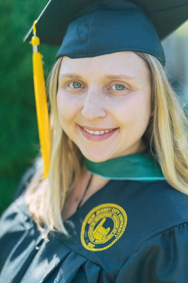 A young woman in a graduation cap and gown smiling outdoors, with a green background.