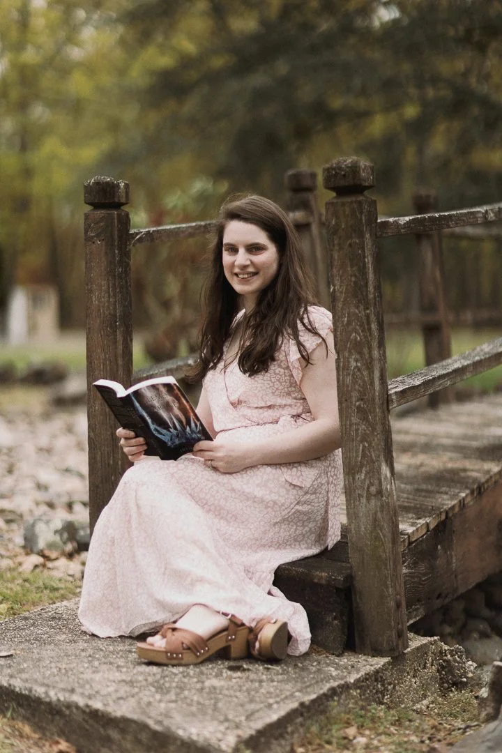 A young woman with long brown hair sitting on a wooden bridge outdoors, holding a book, wearing a pink dress and tan sandals, with trees in the background.