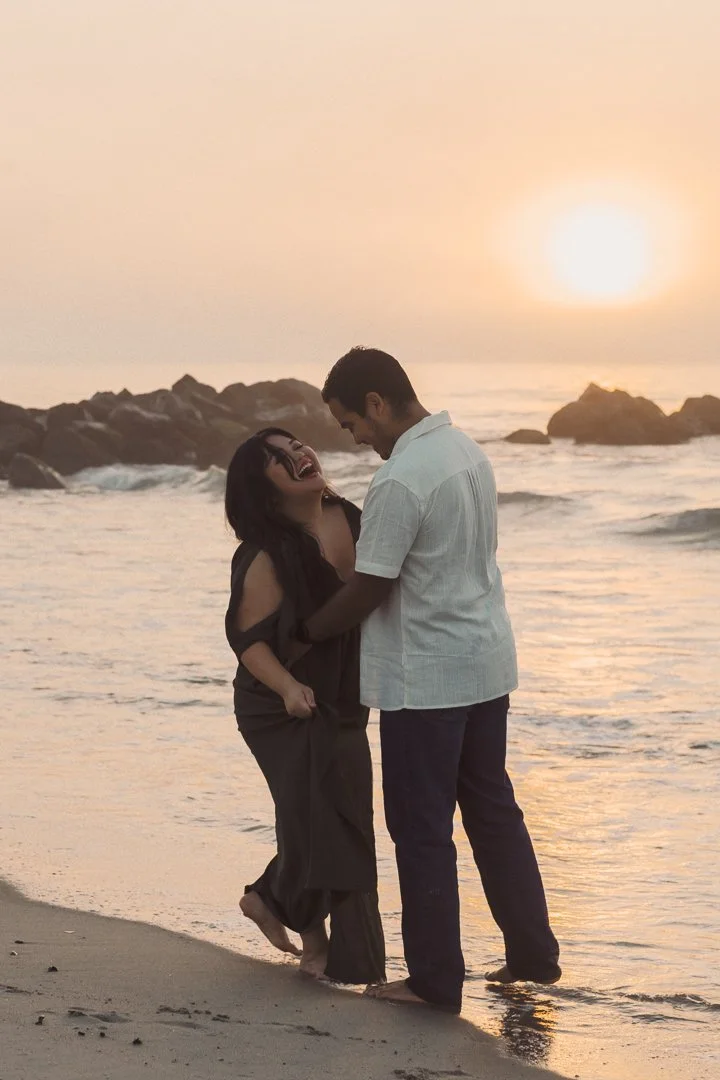 A couple laughs and enjoys each other's company on the beach during sunset, standing close with the ocean and rocks in the background.