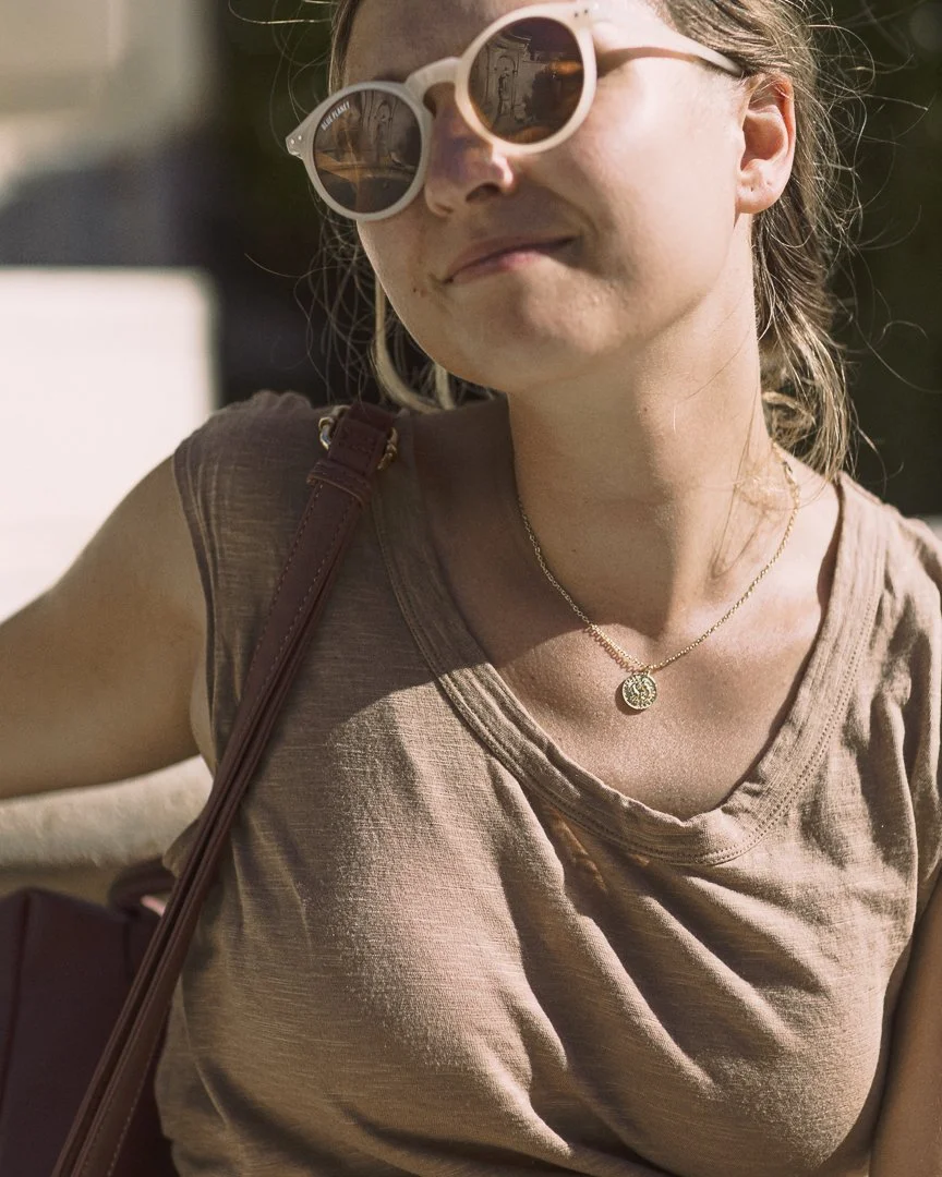 A woman wearing white sunglasses, a beige t-shirt, a gold necklace, and carrying a brown shoulder bag, outdoors in the sunlight.