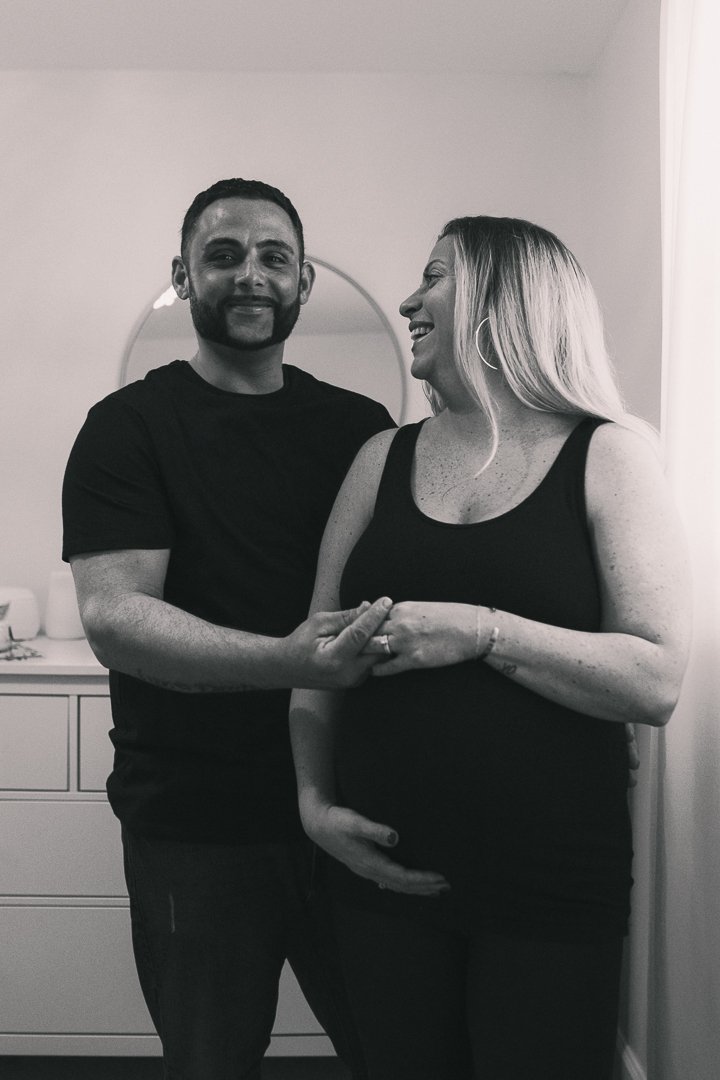A couple standing indoors, with the woman showing a pregnant belly, smiling and looking at each other, in black and white.