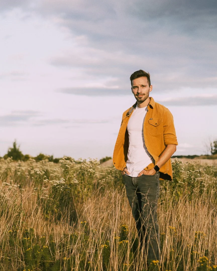 A man in a mustard yellow jacket, white T-shirt, and dark pants standing in a wildflower field under a partly cloudy sky.