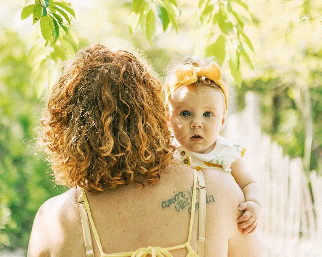 A woman with curly red hair is holding a young girl with blonde hair and a yellow headband outdoors with green leaves in the background.