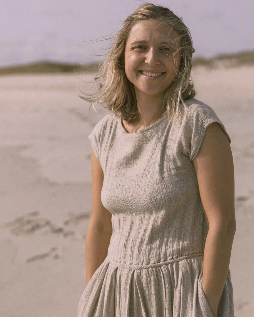 A woman with blonde hair smiling on a sandy beach, wearing a light-colored dress with her hands in pockets.
