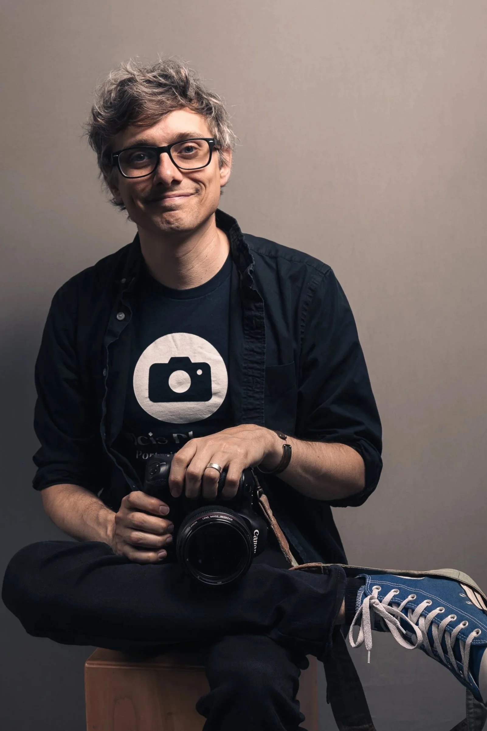 A man with curly hair and glasses, smiling slightly while holding a camera, sitting on a wooden stool against a plain wall. He wears a black shirt with a camera logo, jeans, and sneakers. He is lounging with one leg crossed over his knee.