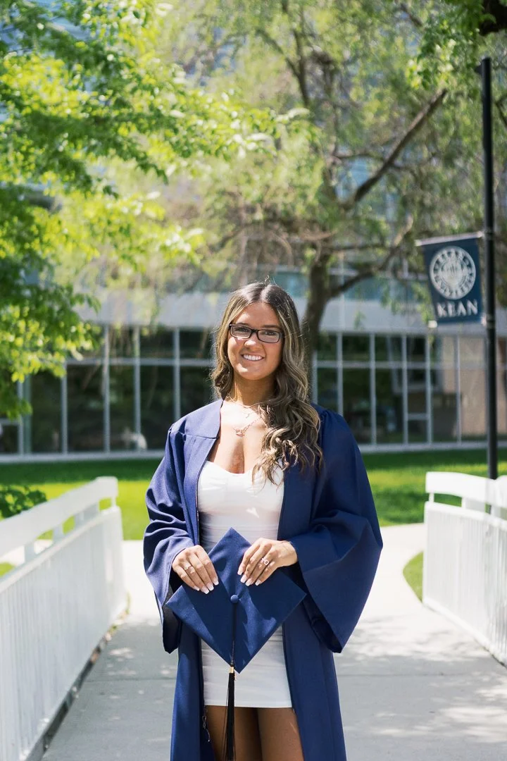 A young woman in a blue graduation gown holding a cap, standing outdoors on a sunny day, smiling for the camera with trees and a building in the background.