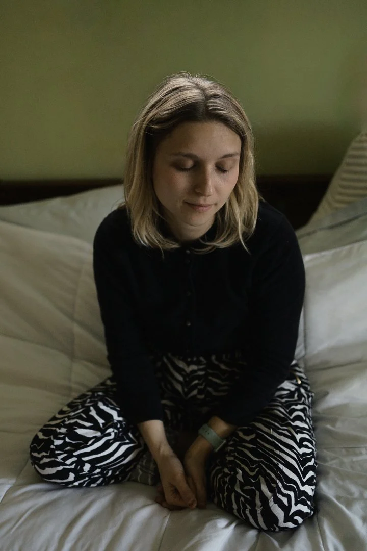 A woman with shoulder-length blonde hair sitting cross-legged on a bed, eyes closed, wearing a black shirt and zebra-print pajama pants, with a green wall in the background.