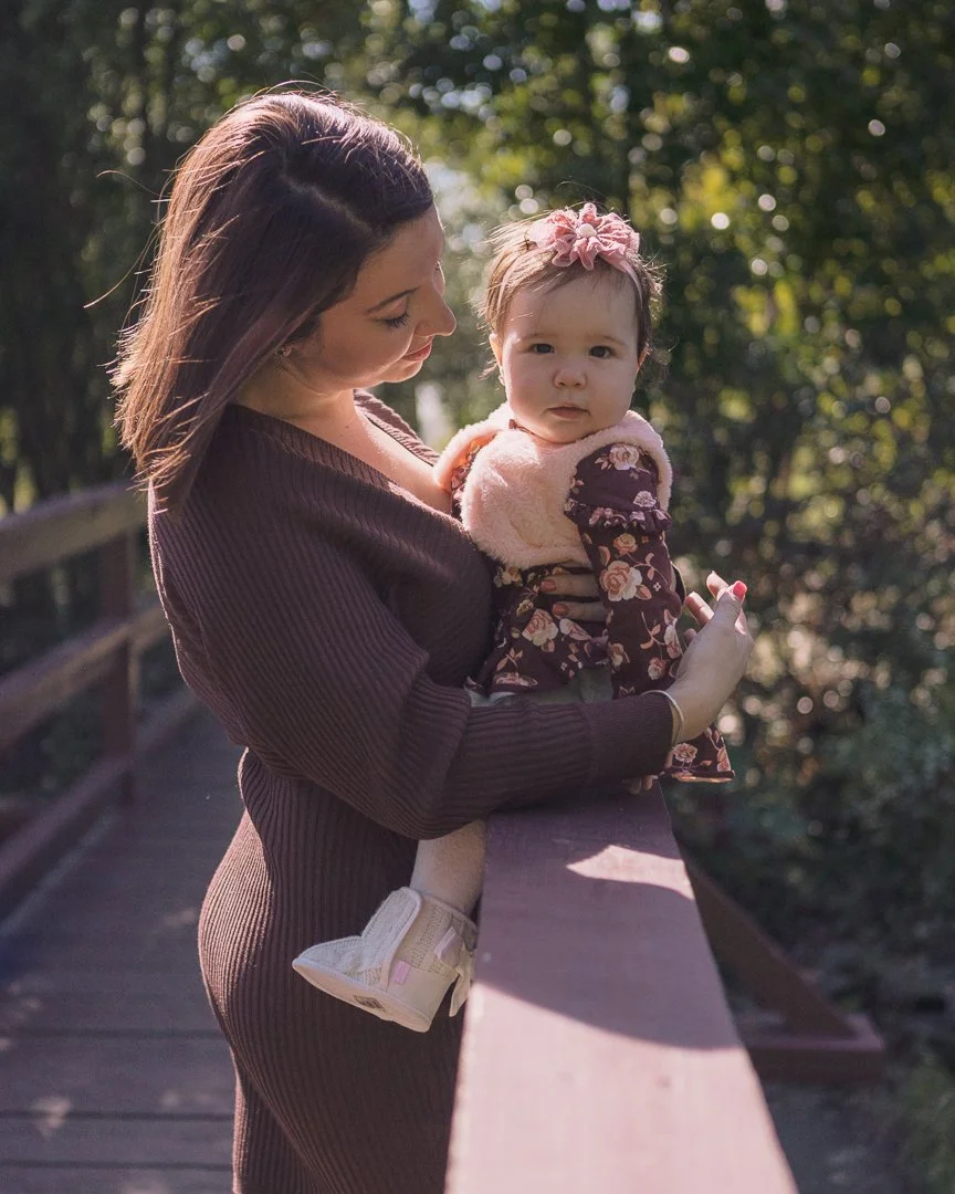 A woman holding a young girl on a wooden bridge outdoors surrounded by greenery.