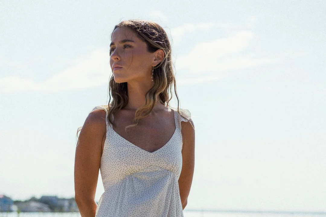 A young woman with wavy brown hair wearing a white polka-dot sleeveless dress, standing outdoors near water, with a cloudy sky behind her.