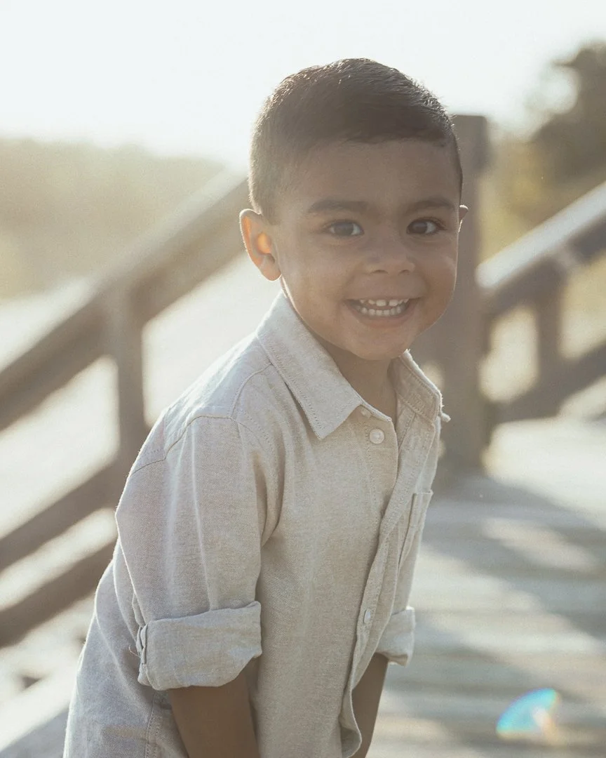 A young boy with short dark hair wearing a light-colored button-up shirt, smiling and leaning against a wooden railing outdoors with sunlight and a blurred background.