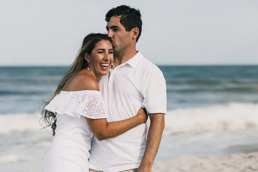 A couple hugging and smiling on the beach with ocean waves in the background, woman wearing a white off-shoulder dress and man in a white shirt.