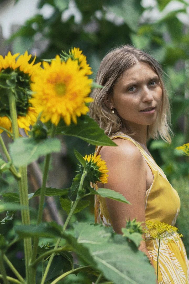 A woman wearing a yellow dress standing among sunflowers in a garden or greenhouse.