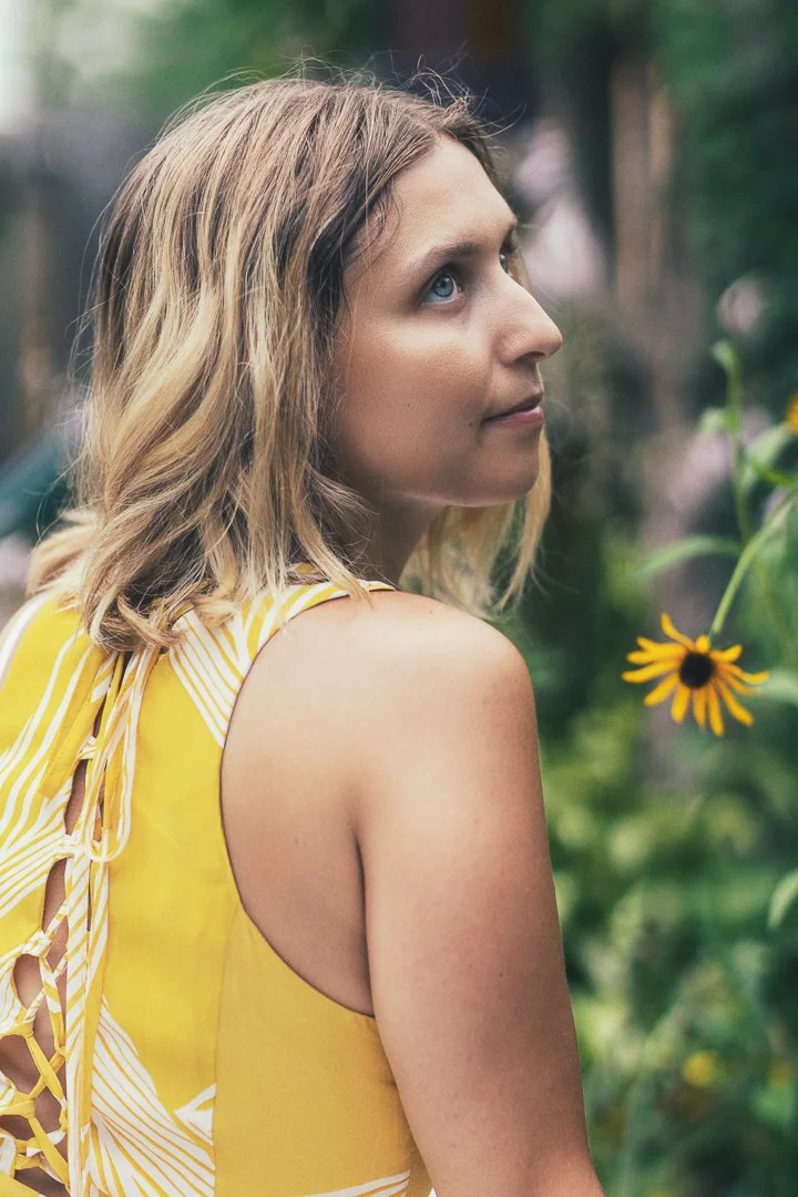 A woman with wavy blonde hair and blue eyes looking sideways, wearing a yellow sleeveless top with lace-up details on the back, outdoors with blurred greenery and a sunflower in the background.