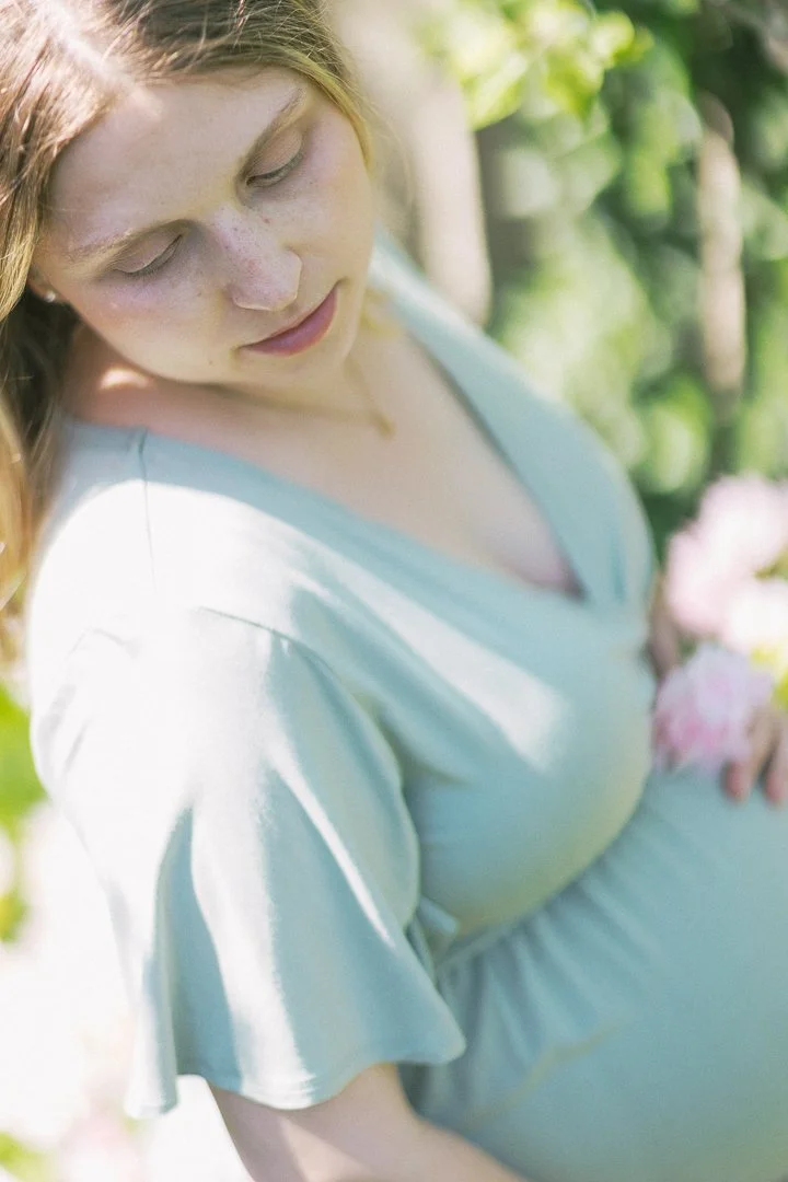 A pregnant woman with red hair in a light blue dress holding pink flowers outdoors, with greenery in the background.