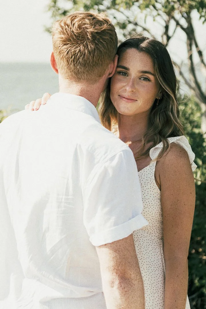 A woman with brown hair and a light-colored dress smiling at the camera while a man with blond hair, wearing a white shirt, has his back to the camera and leans in close to her outdoors near trees and water.