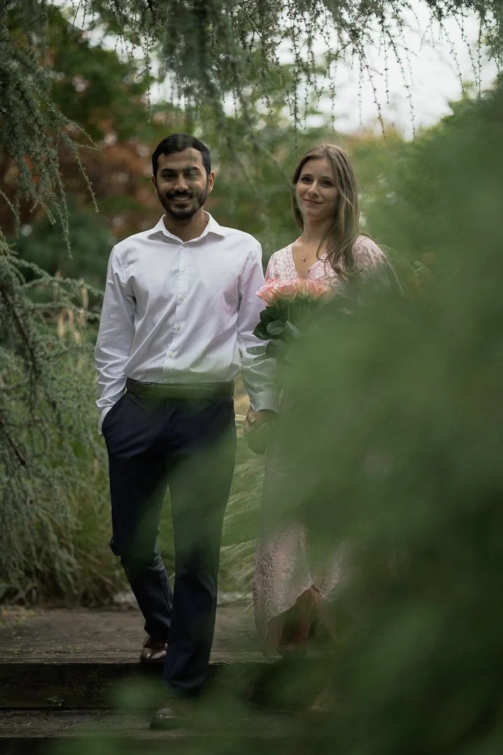 A man and woman walking hand in hand on a trail in a wooded area. The man has dark hair and a beard, wearing a white shirt and dark pants. The woman has light brown hair, wearing a patterned dress, and holding a bouquet of pink roses. They are smilin