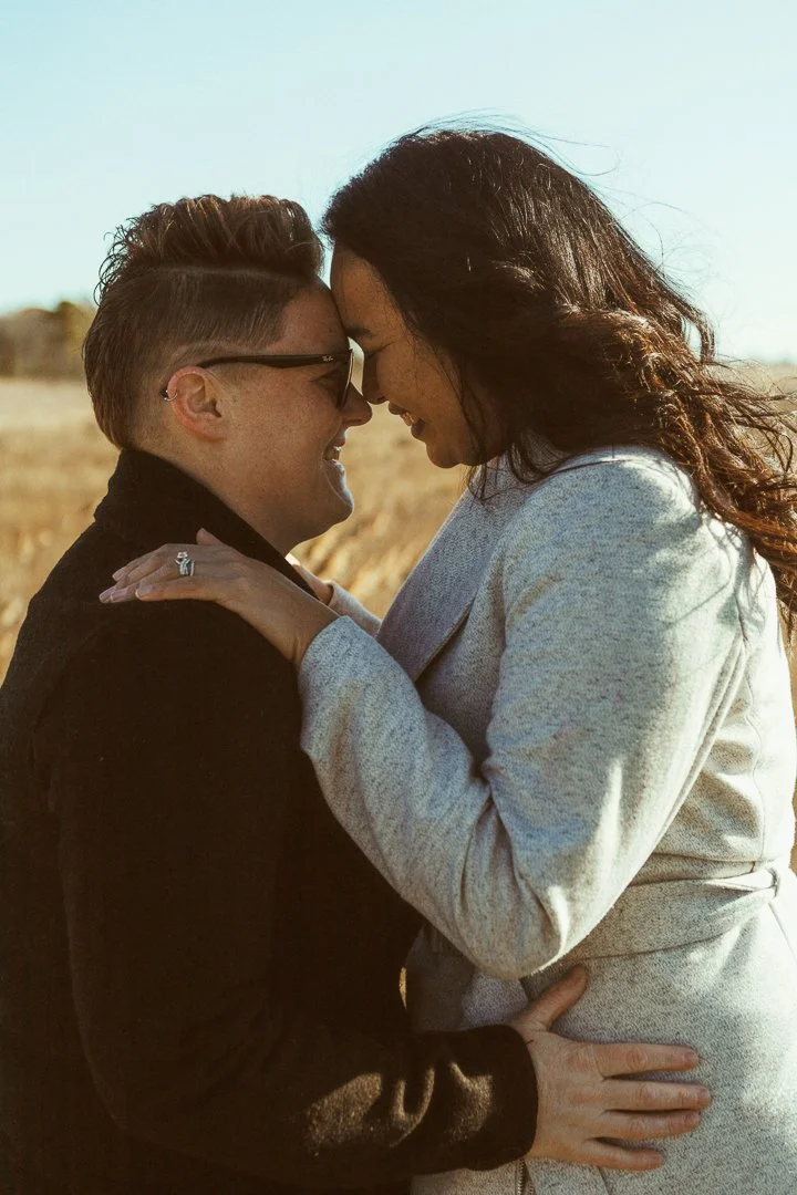 Couple embracing outdoors in a field, touching foreheads and smiling, with a clear sky in the background.