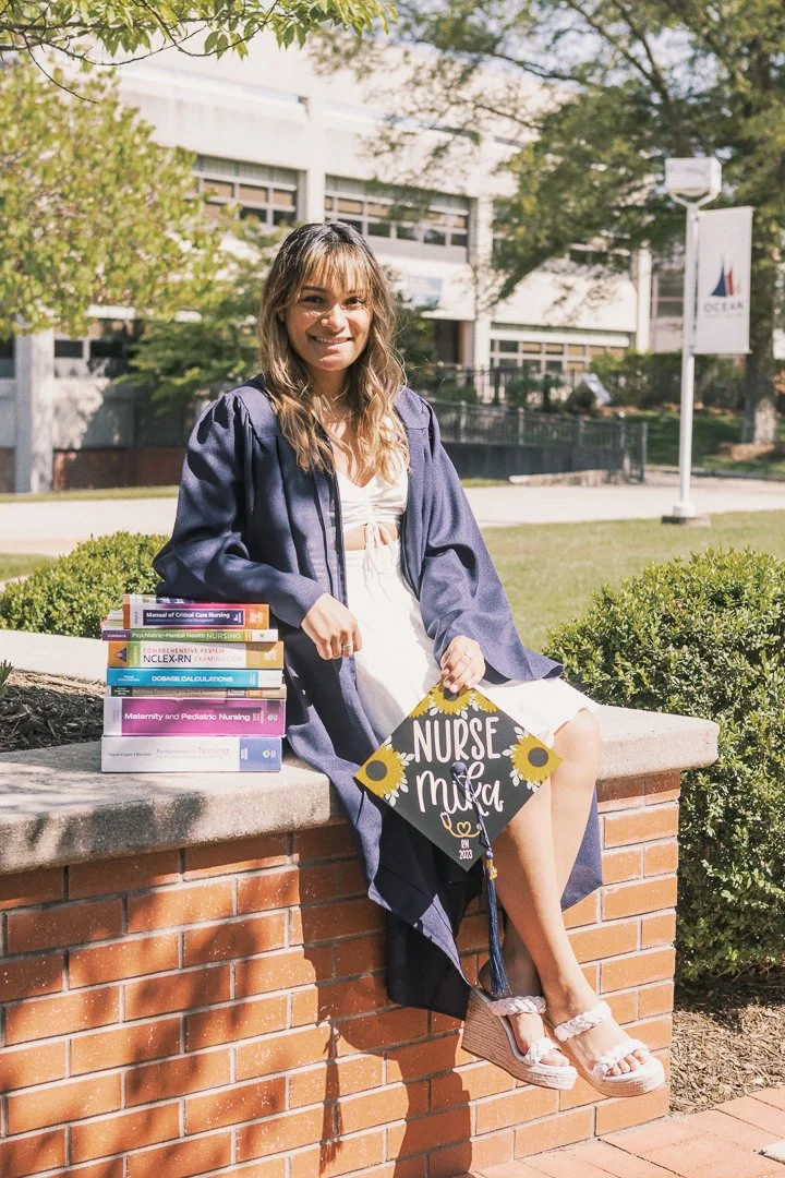 Young woman in graduation gown sitting on a brick wall outdoors, holding a decorated graduation cap that reads 'Nurse Mia' with sunflower designs, with a stack of nursing textbooks beside her, smiling on her graduation day.