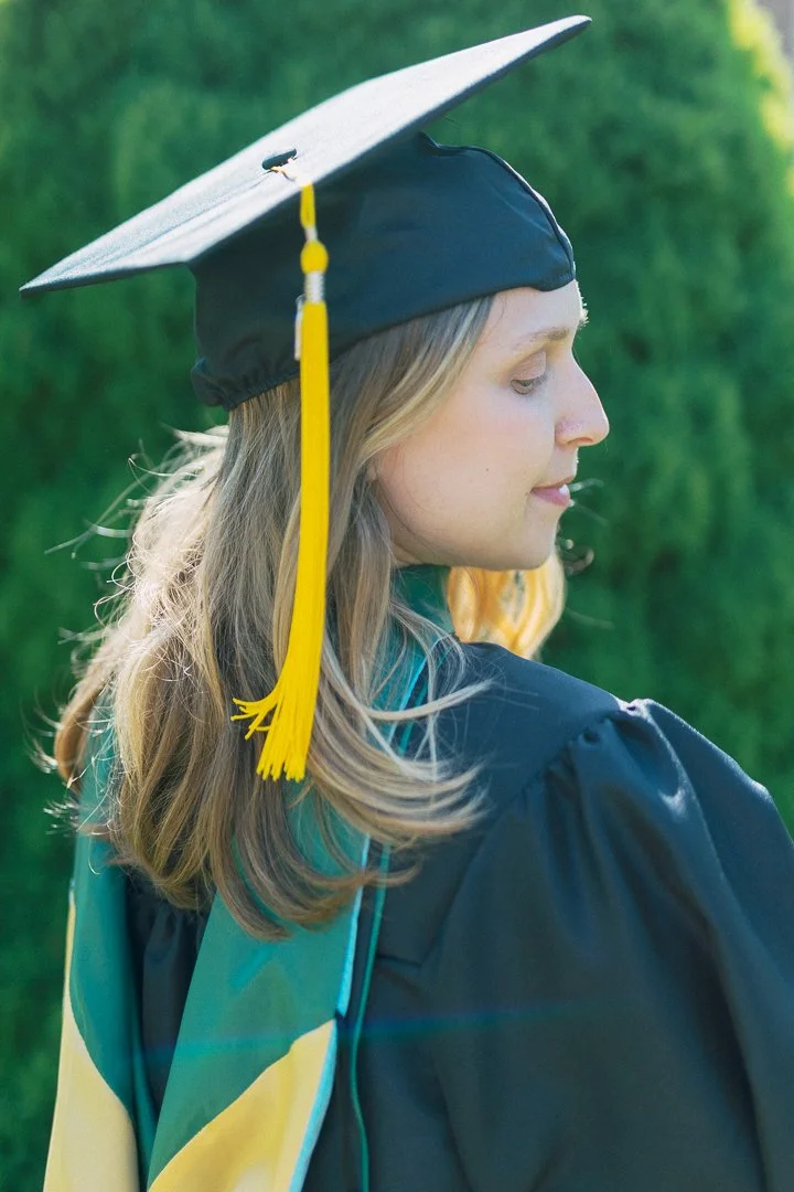 A young woman with long blonde hair in a cap and gown, standing outdoors with a blurred green background.