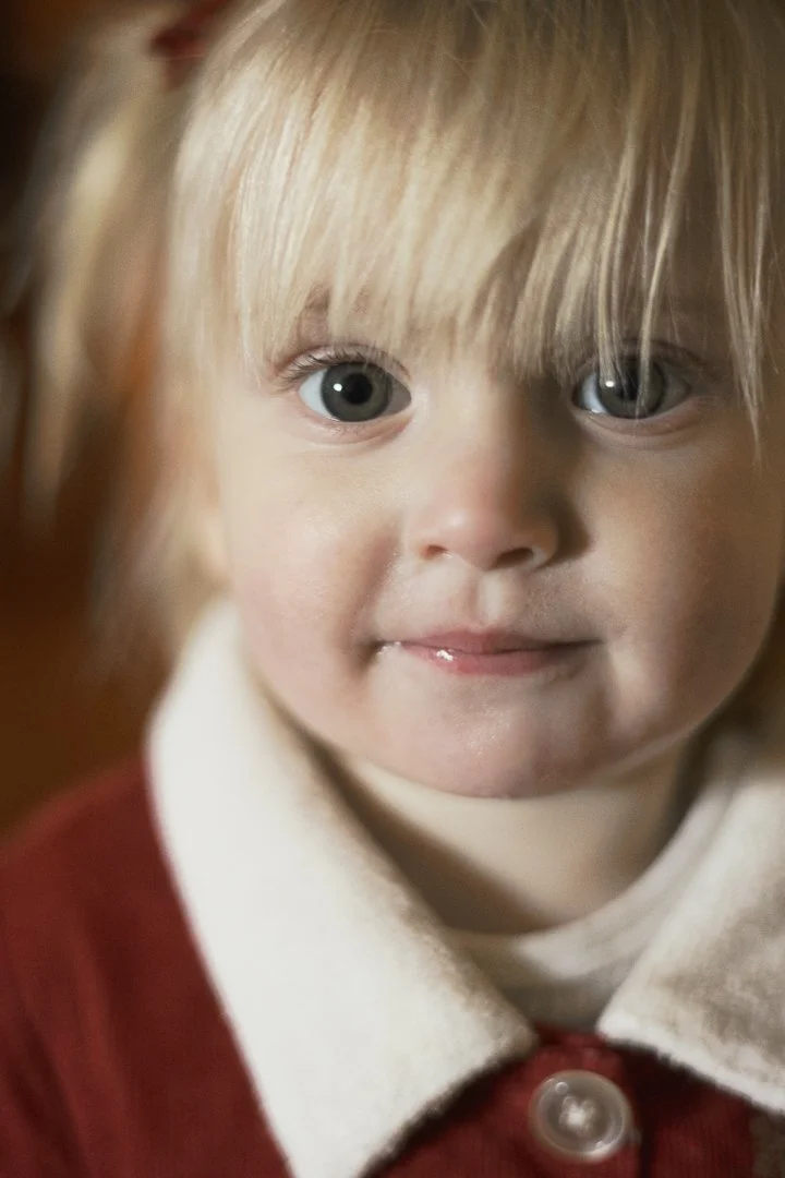 Close-up of a young girl with blonde hair and big gray eyes, wearing a red coat with a buttoned white collar.
