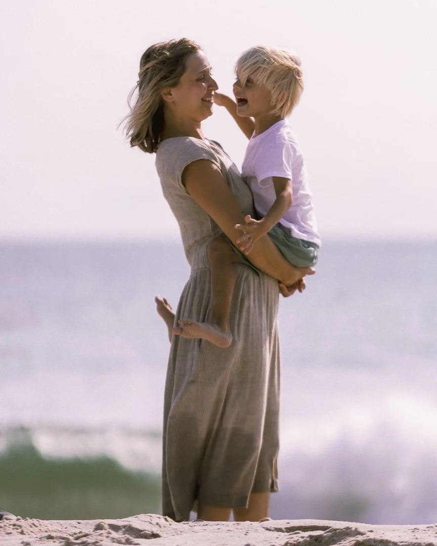 A woman holding a young boy by the beach, smiling and looking at each other with the ocean in the background.