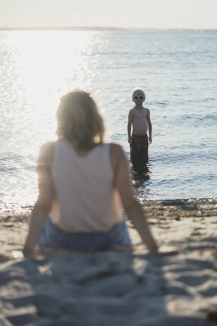 A woman sitting on the beach, watching a boy in sunglasses standing in the water at sunset or sunrise.