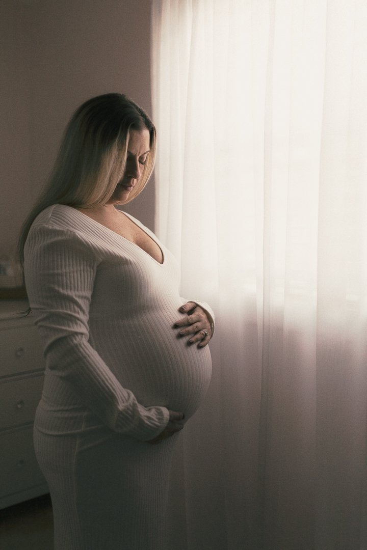 Pregnant woman with blonde hair standing near a window with sheer curtains, wearing a white long-sleeve top and matching pants, gently touching her belly.