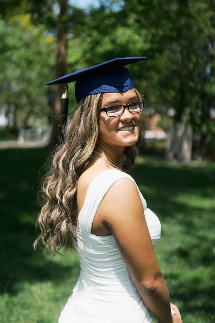 A young woman with long wavy hair, wearing glasses and a white dress, standing outdoors on a sunny day, smiling and wearing a graduation cap.