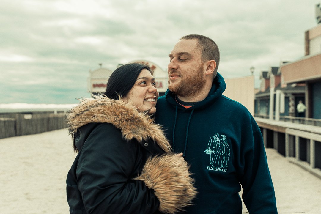 A couple standing close together outdoors near a beach, with the woman smiling at the man who is looking at her, both dressed in warm clothing.