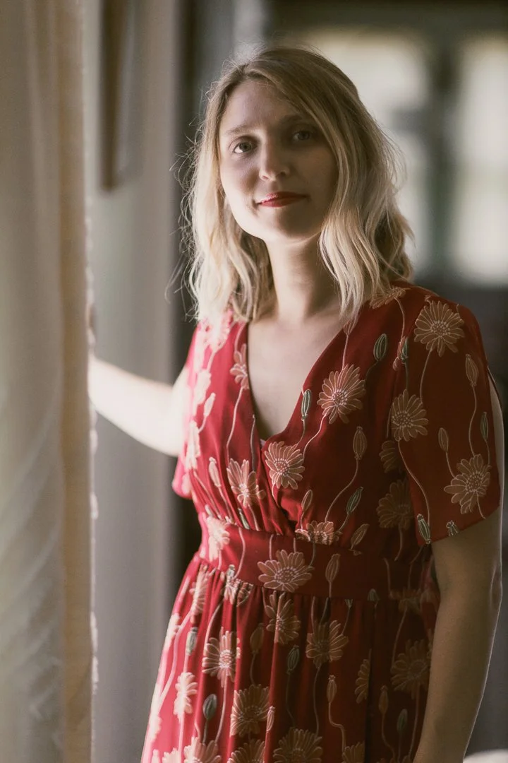 A woman with shoulder-length blonde hair wearing a red dress with pink floral patterns, standing near a window with soft natural light