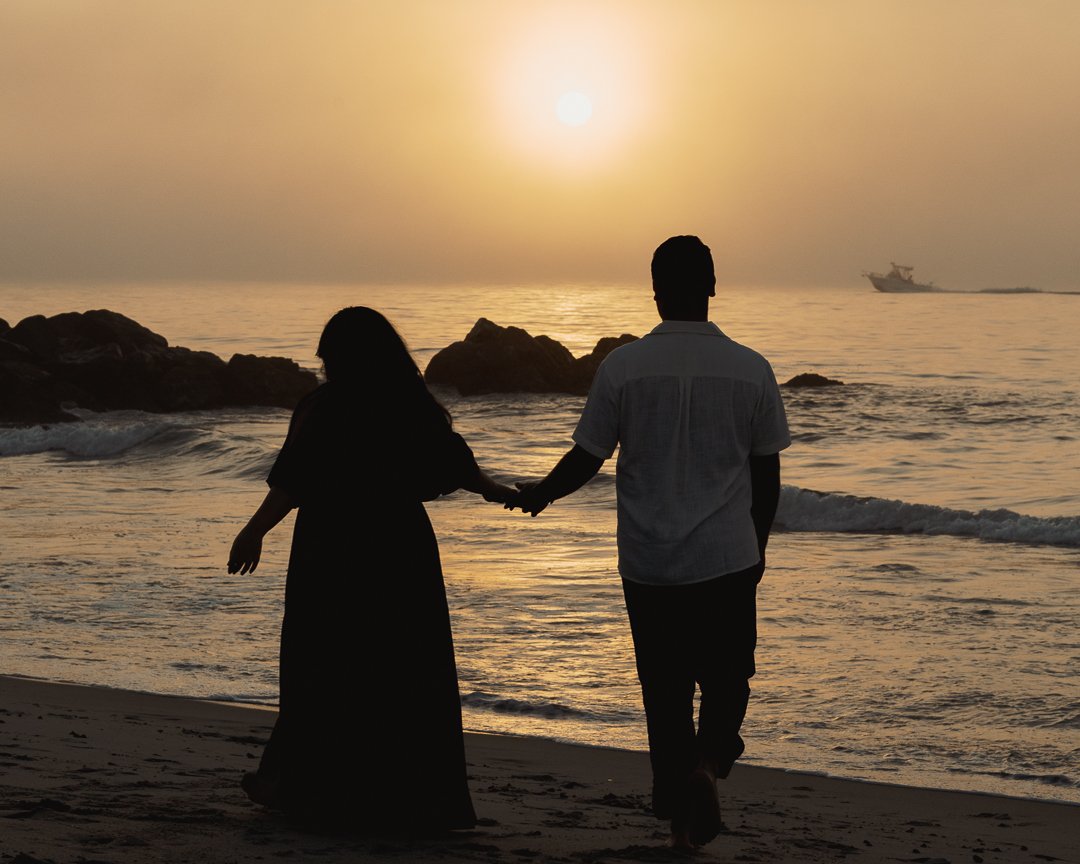 Silhouette of a couple holding hands and walking along a beach at sunset.
