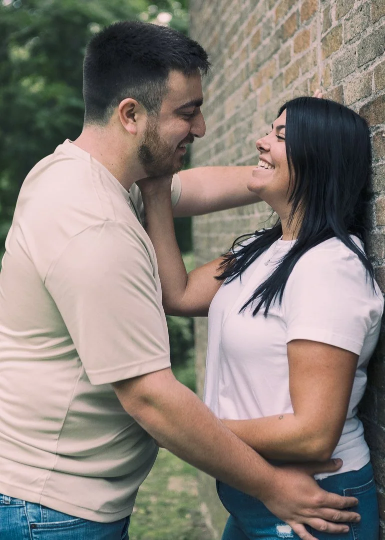 A couple smiling and looking at each other with their faces close, outside near a brick wall and greenery.
