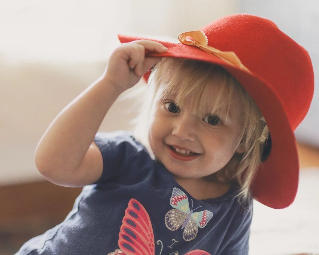 A young girl with blonde hair, blue eyes, and a big smile, wearing a large red floppy hat with a bow and a navy blue shirt with a butterfly patch, tilting her head and holding the hat with her right hand.