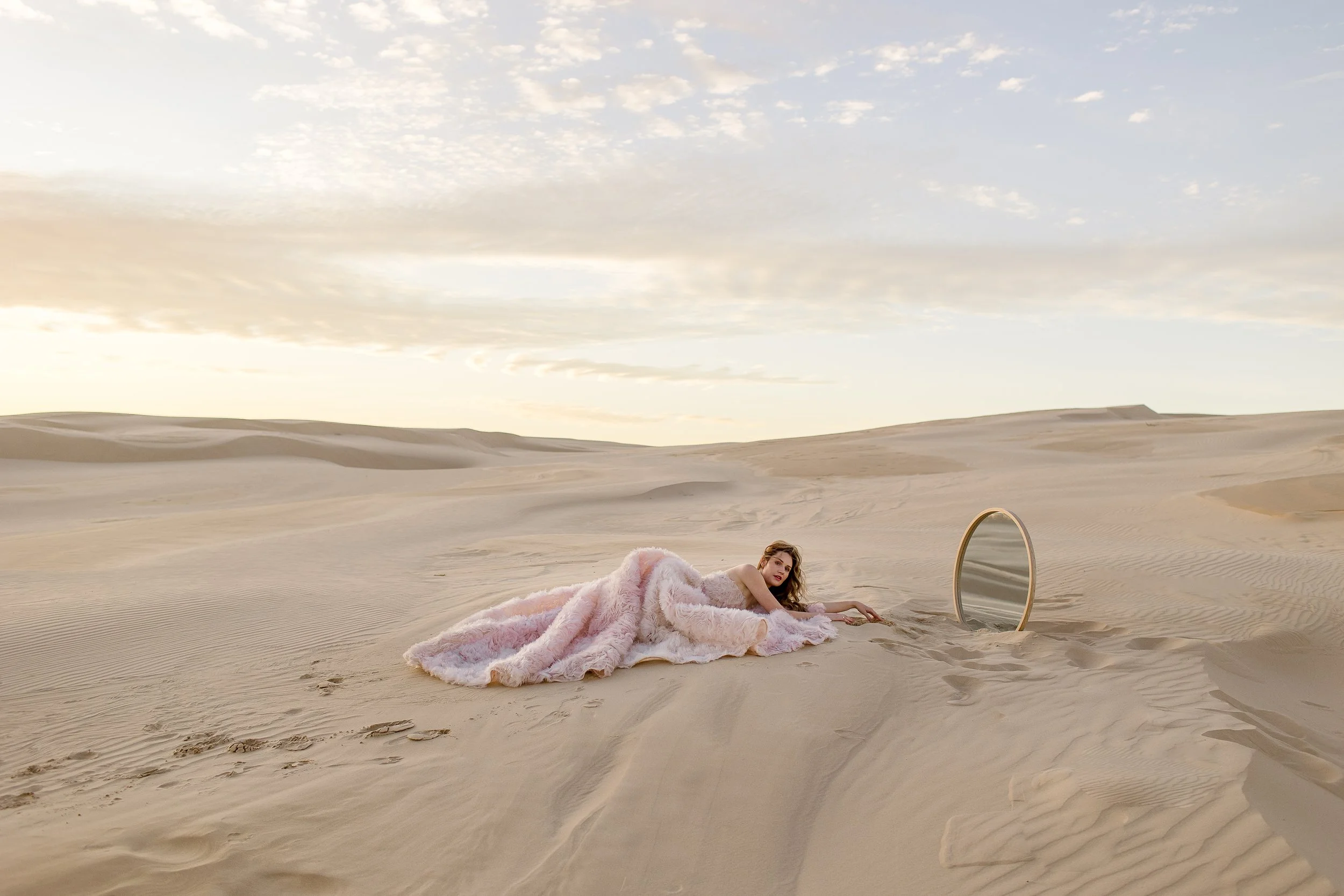 A woman lying on the sand in a desert landscape, covered with a pink fluffy blanket, near a standing mirror reflecting the sky.