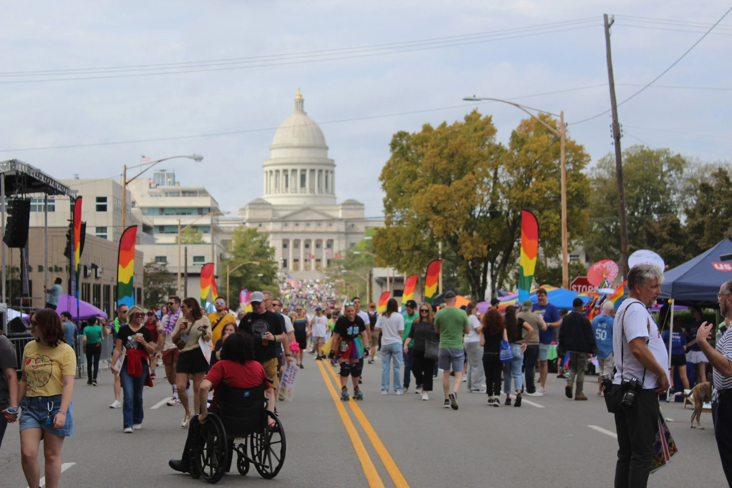 A street filled with people attending an outdoor event, with colorful flags, tents, and a white capitol building in the background.