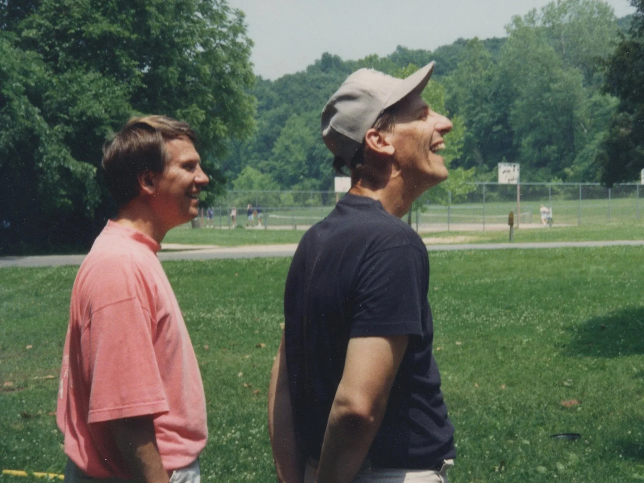 Two men smiling and standing outdoors on a grassy area, with trees and a park in the background.