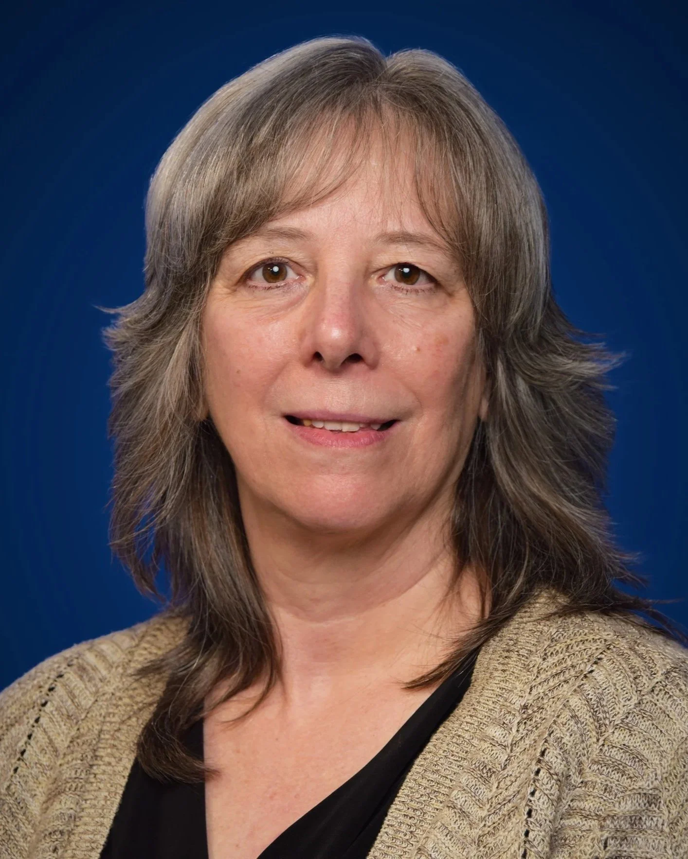 A woman with shoulder-length gray hair wearing a beige knitted sweater and black top, posing against a blue background.
