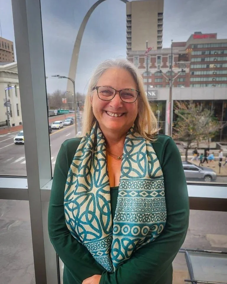 A smiling woman with glasses and gray hair standing indoors near a large window, with the Gateway Arch and city buildings visible outside.