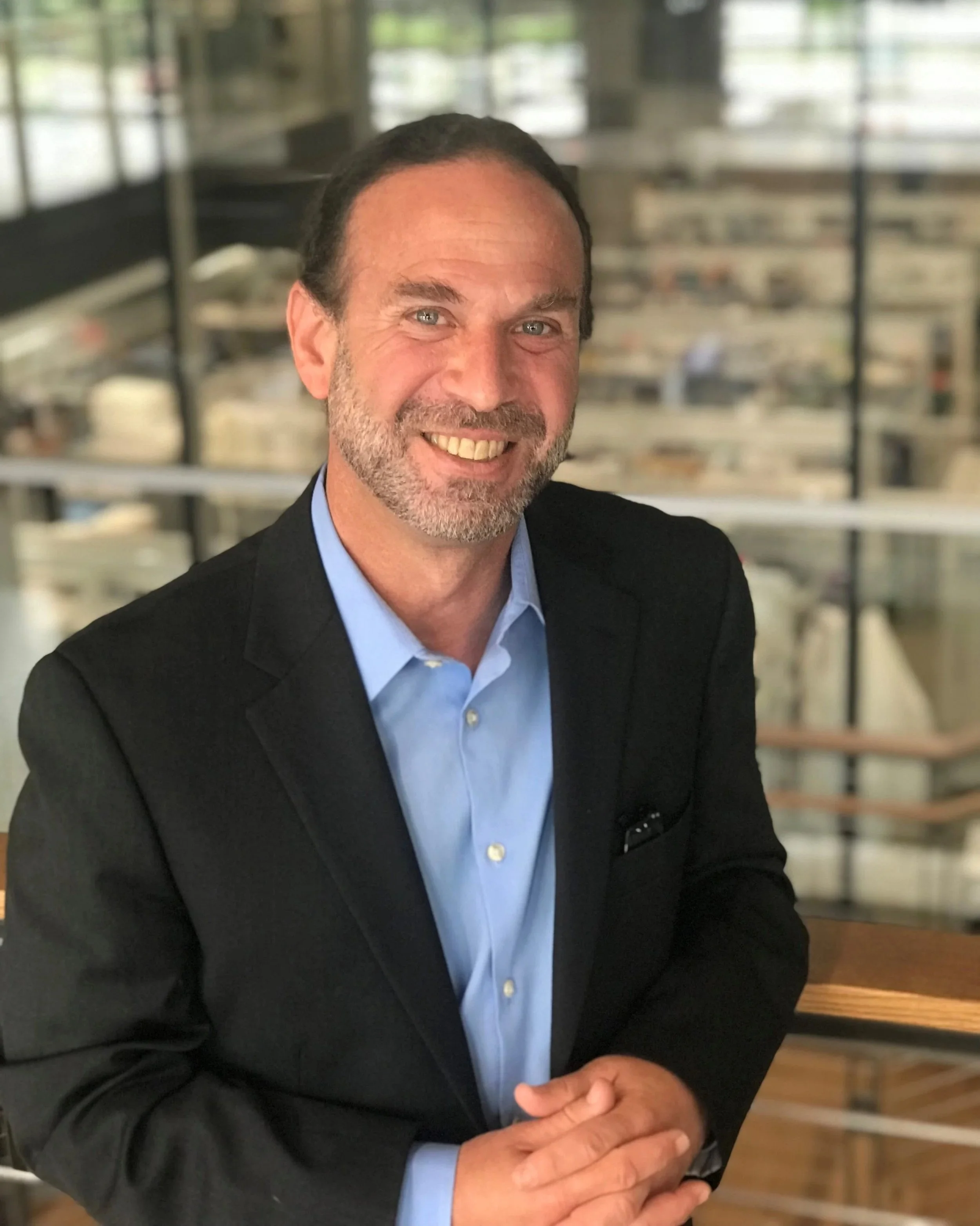 A man with dark hair and a beard, wearing a black suit jacket and a light blue shirt, smiling and standing indoors with a blurred background of glass walls and bookshelves.