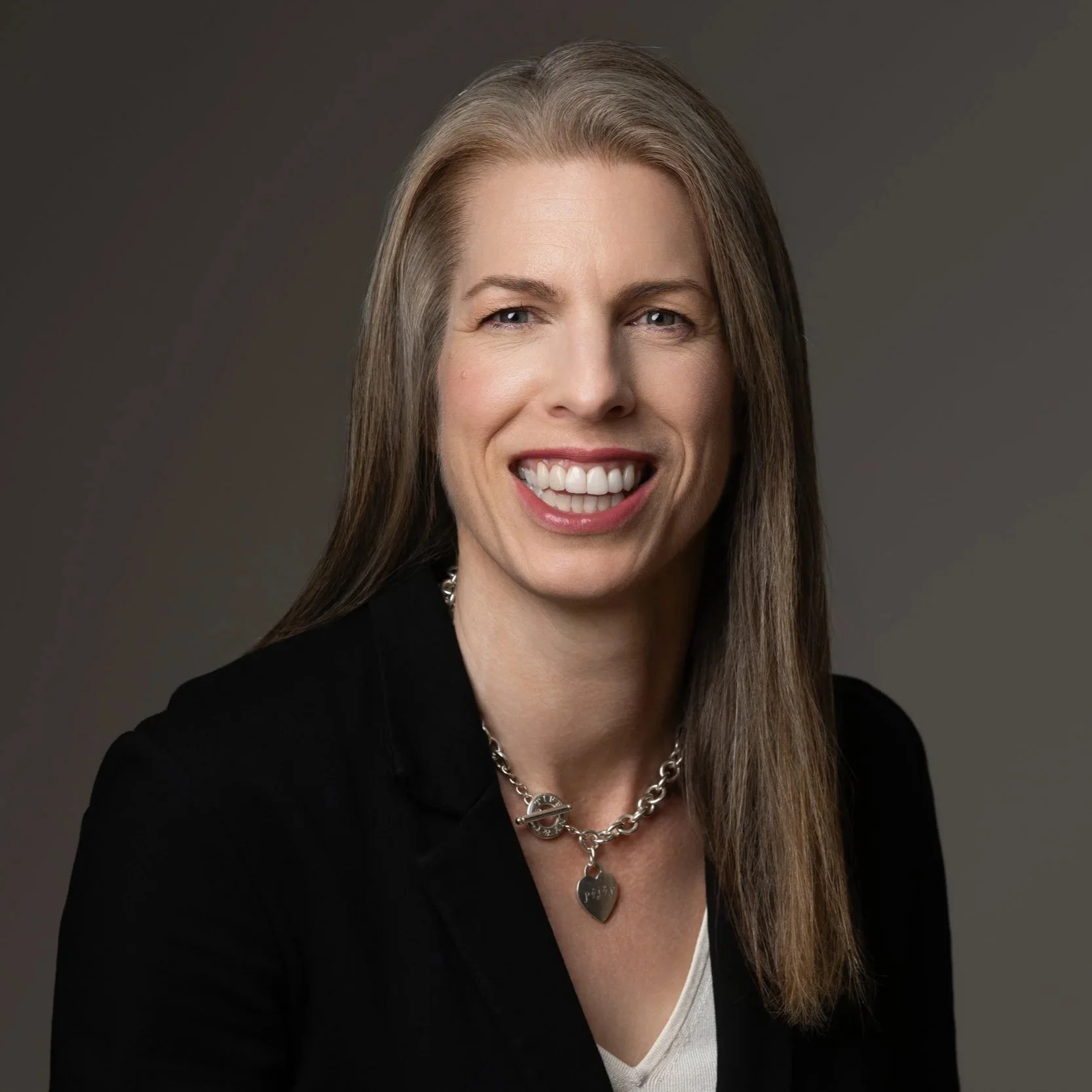 Jenn Lofgren MCC wearing a black top, silver necklaces, and pink lipstick, smiling in front of a blurred red brick wall background.