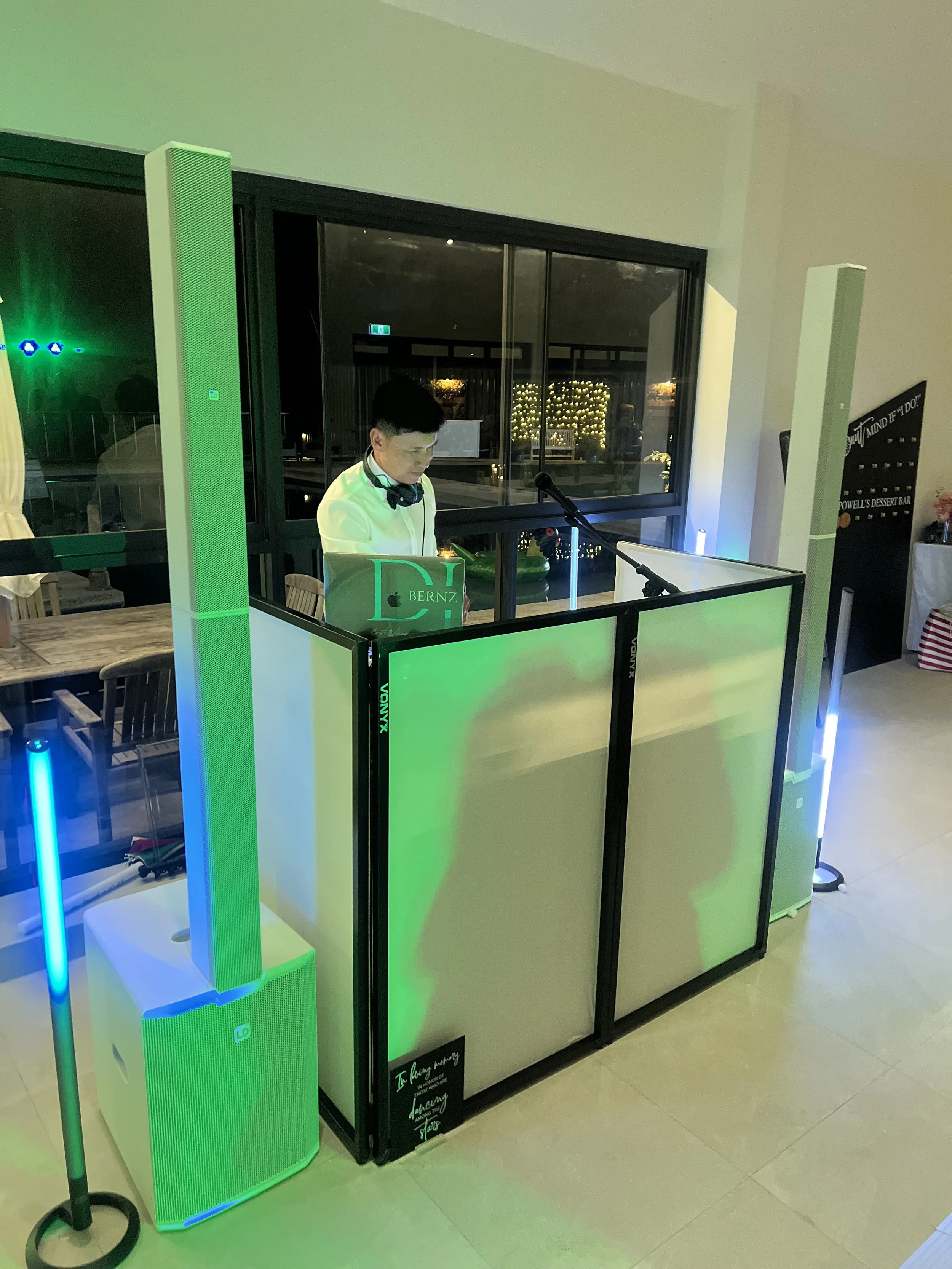 A person wearing headphones and a white shirt DJs at a small booth with green lighting, in a room with large window at night showing outside decorated with string lights.