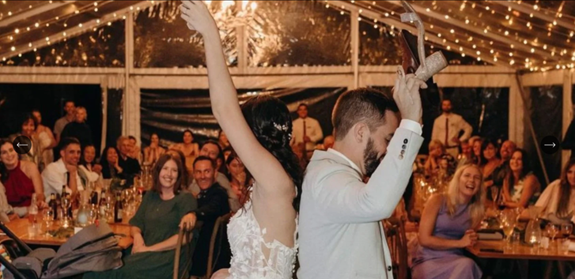 Couple dancing at a wedding reception under string lights with guests sitting at tables in the background.