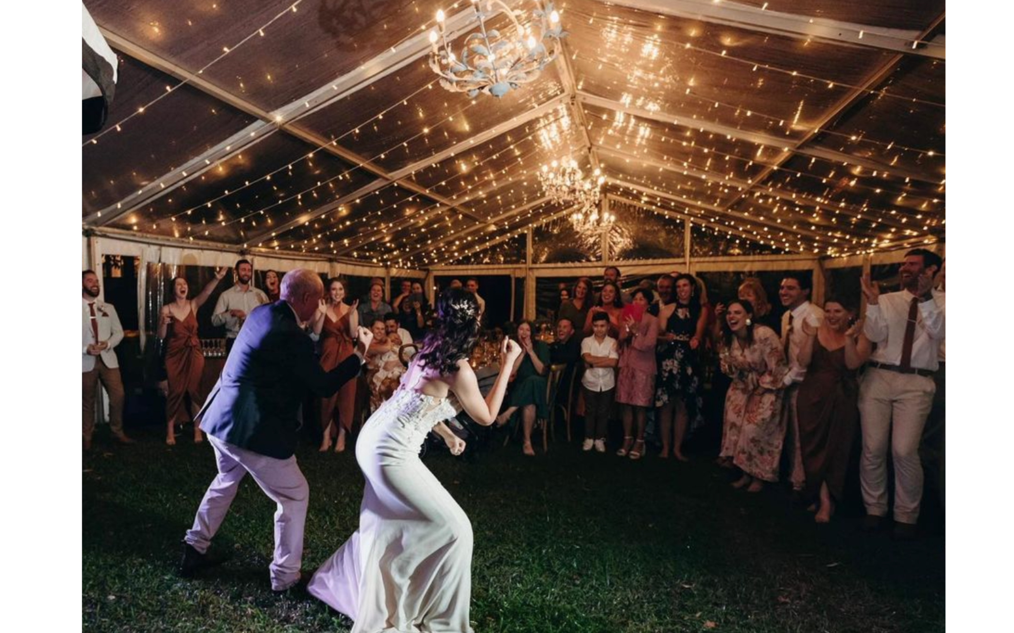 Couple dancing under a tent decorated with string lights at a wedding reception, with guests cheering and laughing in the background.