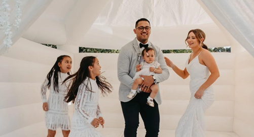 A family at a wedding, with a man holding a baby and a woman in a white dress talking to him, surrounded by three young girls in white dresses.