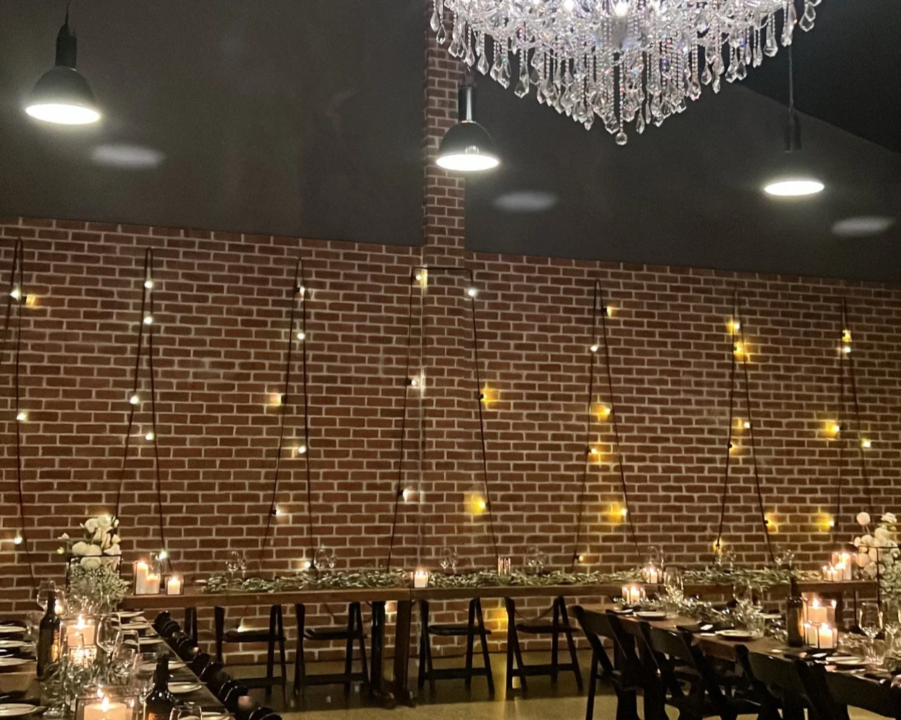 Elegant event space with a long table decorated with candles, flowers, and silverware, against a brick wall. A large crystal chandelier hangs from the ceiling, and string lights are arranged along the wall creating a warm ambiance.
