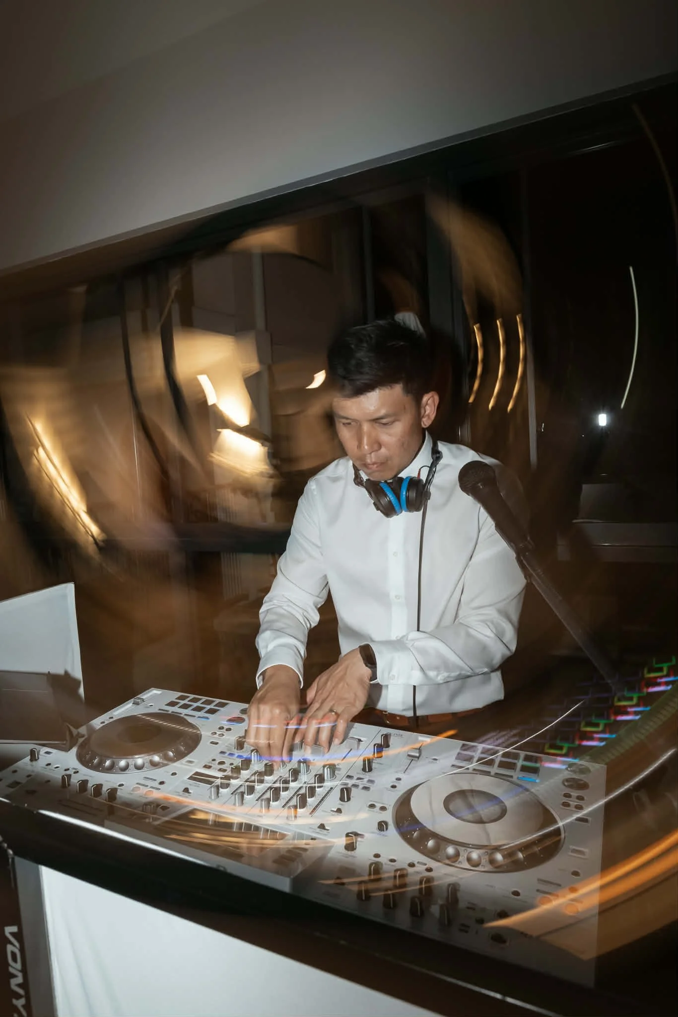 A DJ in a white shirt mixes music at a dark indoor venue, surrounded by DJ equipment and sound controls, with motion blur and swirling lights in the background.