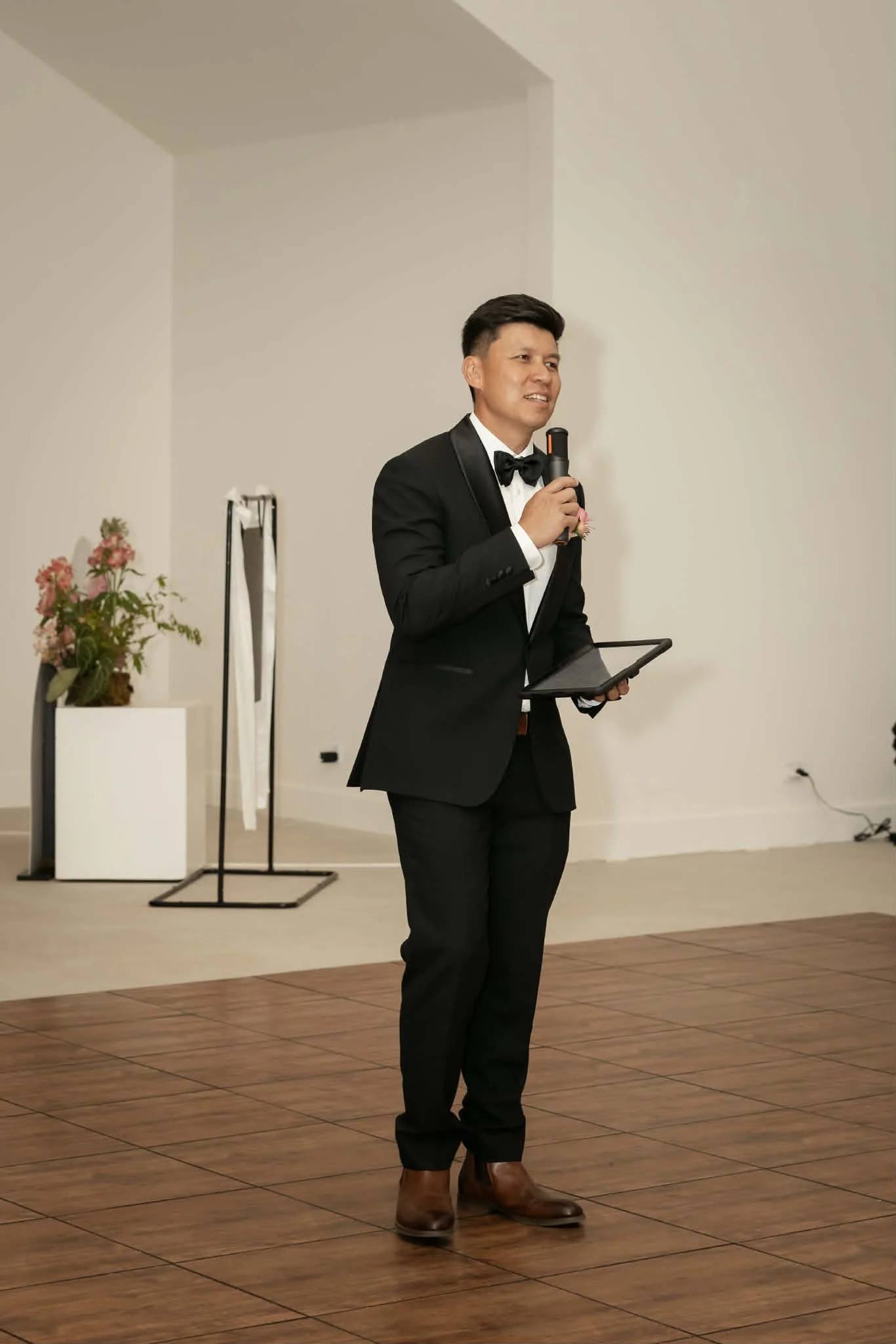 A man in a black tuxedo and bow tie holding a microphone and a tablet, standing on a wooden floor at a formal event.
