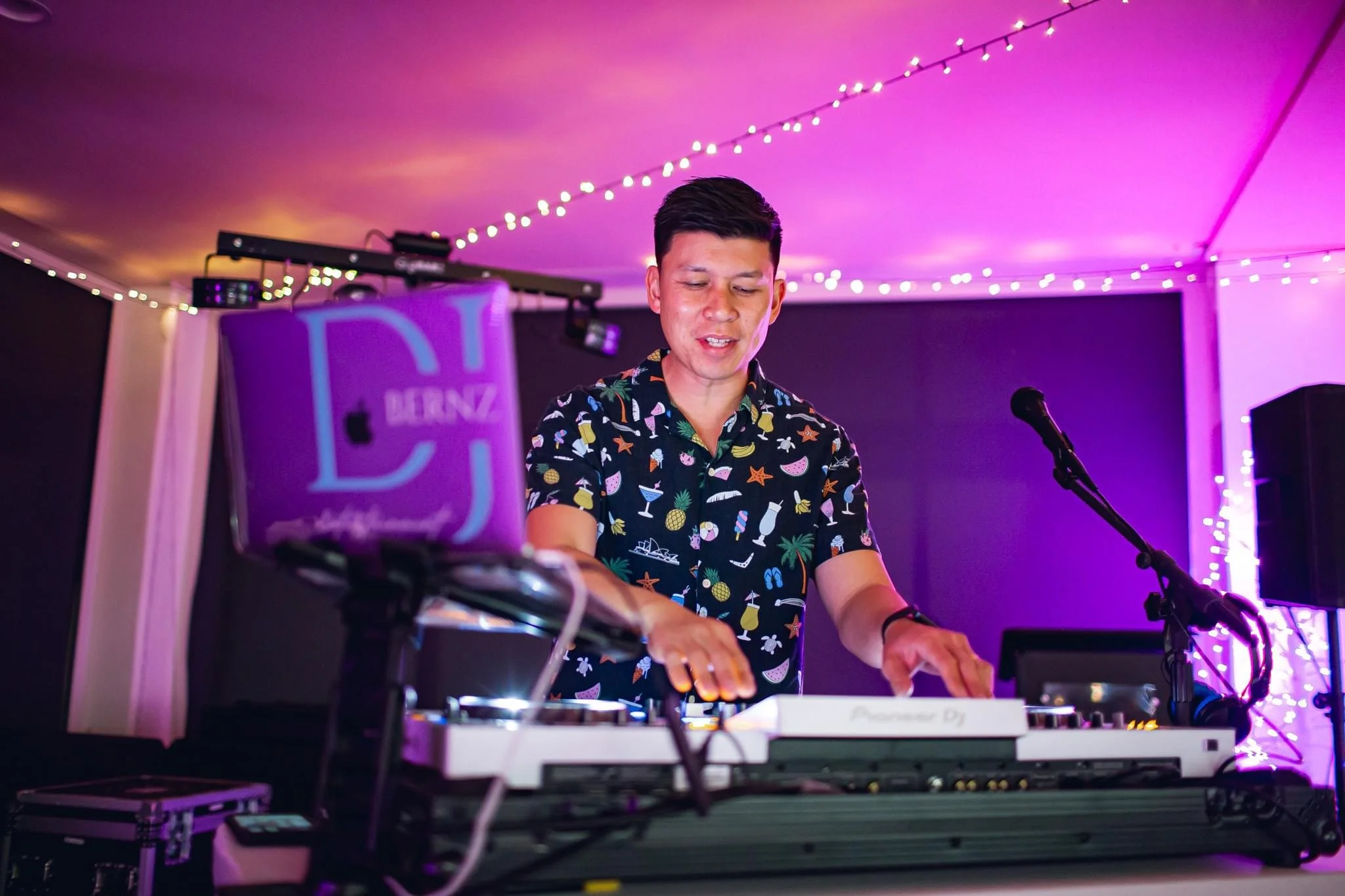 A DJ wearing a colorful tropical shirt mixes music at a party with purple and pink lighting and string lights in the background.
